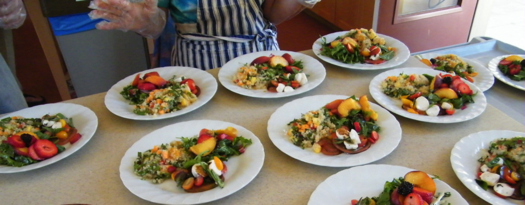 A person in an apron stands over plates of couscous, salad, and fruit